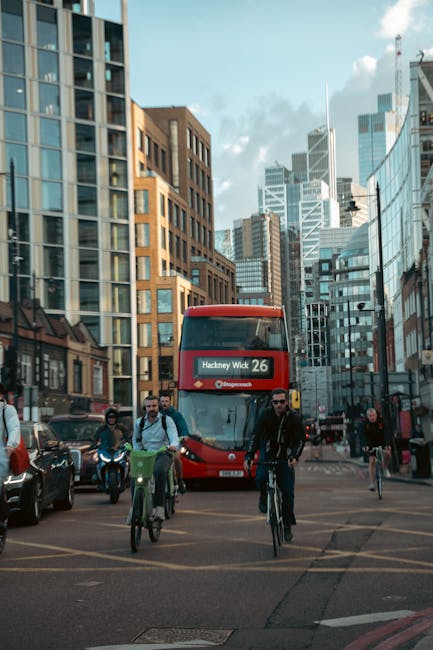 A busy urban street scene in Hackney Wick with modern high-rise buildings and a clear sky in the background. In the foreground, a red double-decker bus displaying 'Hackney Wick 26' on its route sign i
