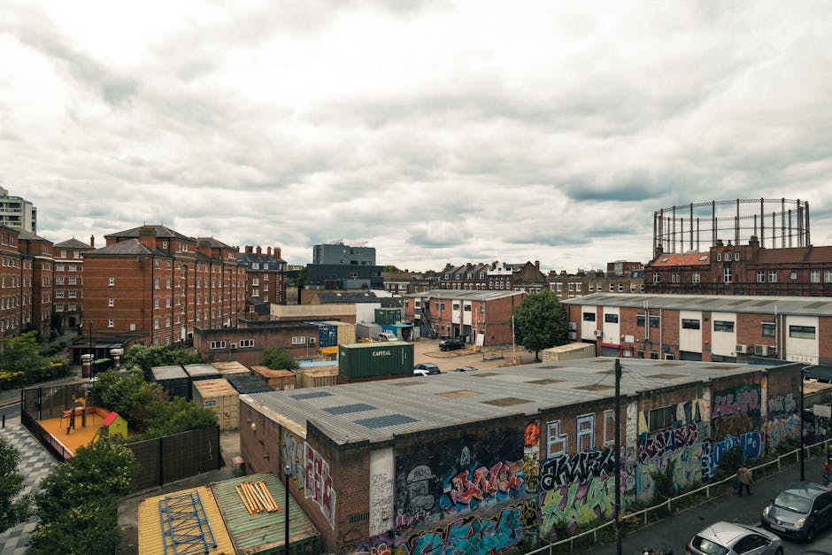 A busy urban street scene in Hackney Wick with modern high-rise buildings and a clear sky in the background. In the foreground, a red double-decker bus displaying 'Hackney Wick 26' on its route sign i