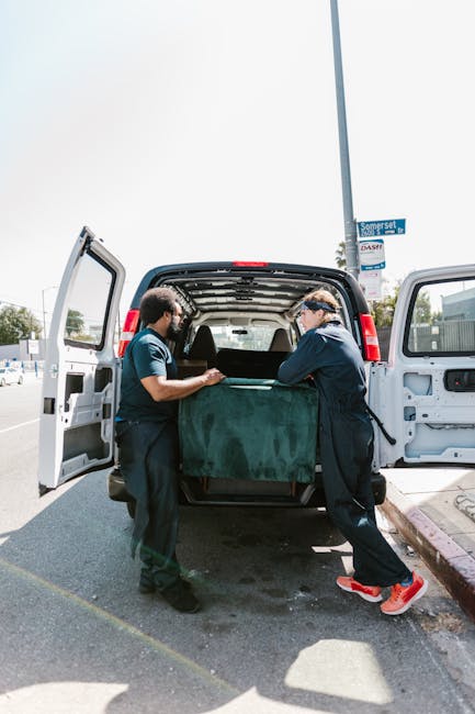 A small flatbed truck loaded with wooden pallets and packaging materials is displayed on a city street, with a white van parked nearby. The truck is positioned in the center of a traffic lane marked w