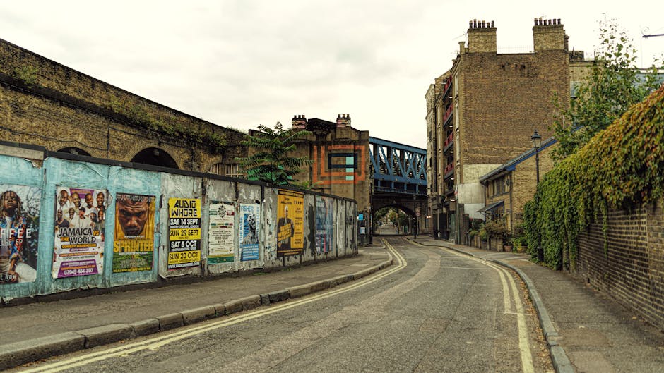 A wide view of the River Lee navigation in Hackney Wick during daylight, with calm water reflecting a partly cloudy sky. On the left side, dense green foliage and overhanging trees partially obscure s
